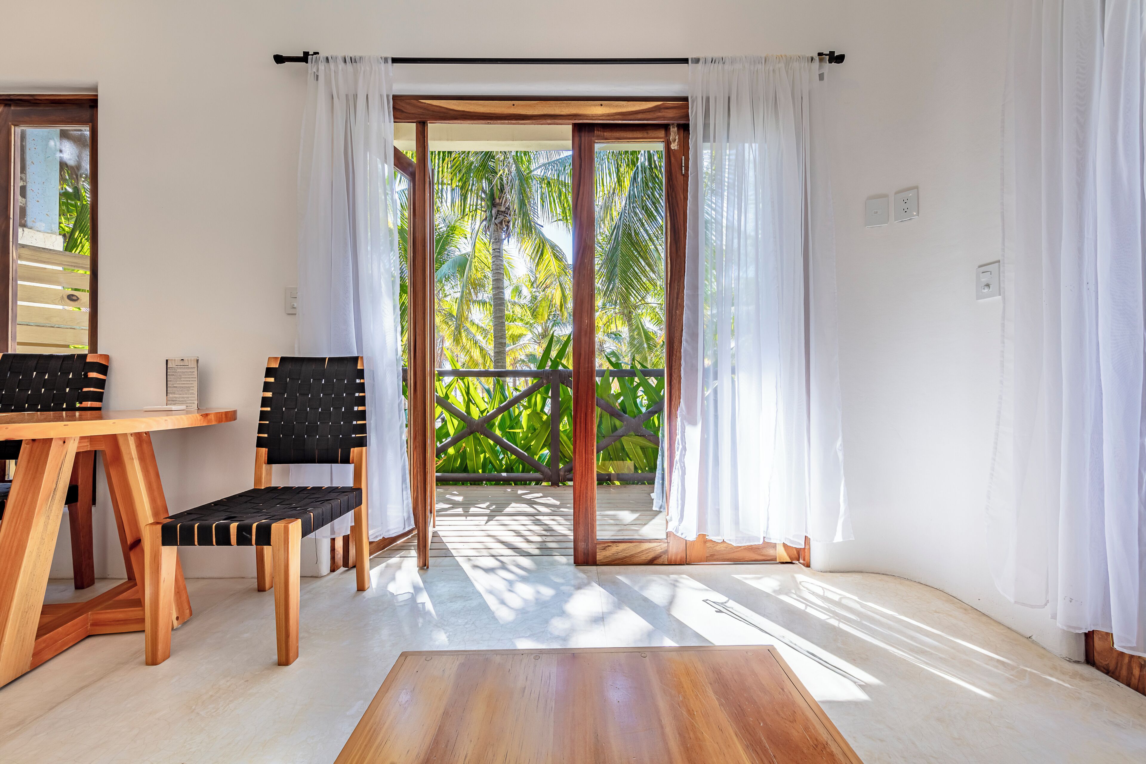 A room with a wooden table, two chairs, a bench, and a view of palm trees through a glass door.