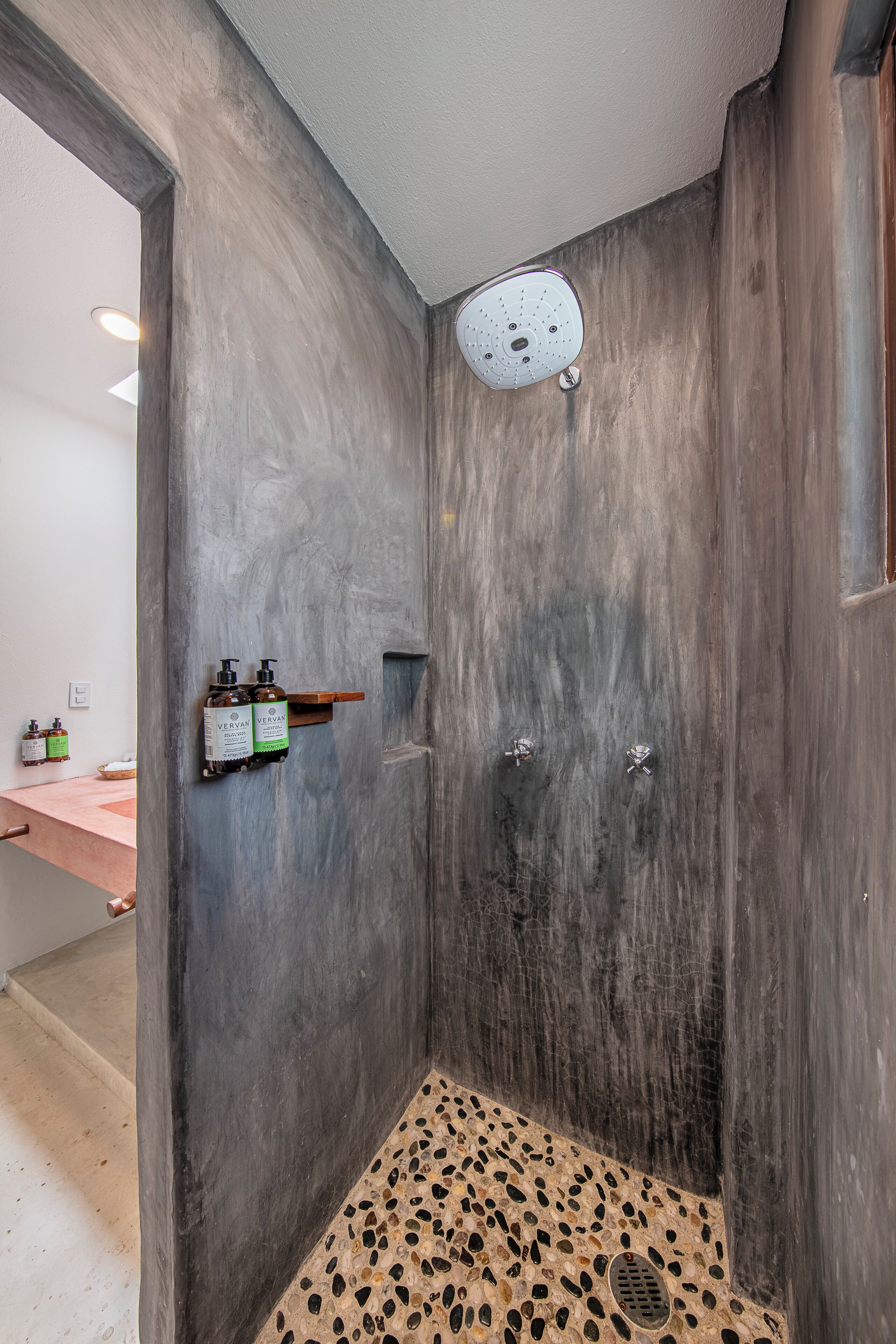 A modern shower with a textured wall, a built-in shelf with toiletries, and a pebble-patterned floor.