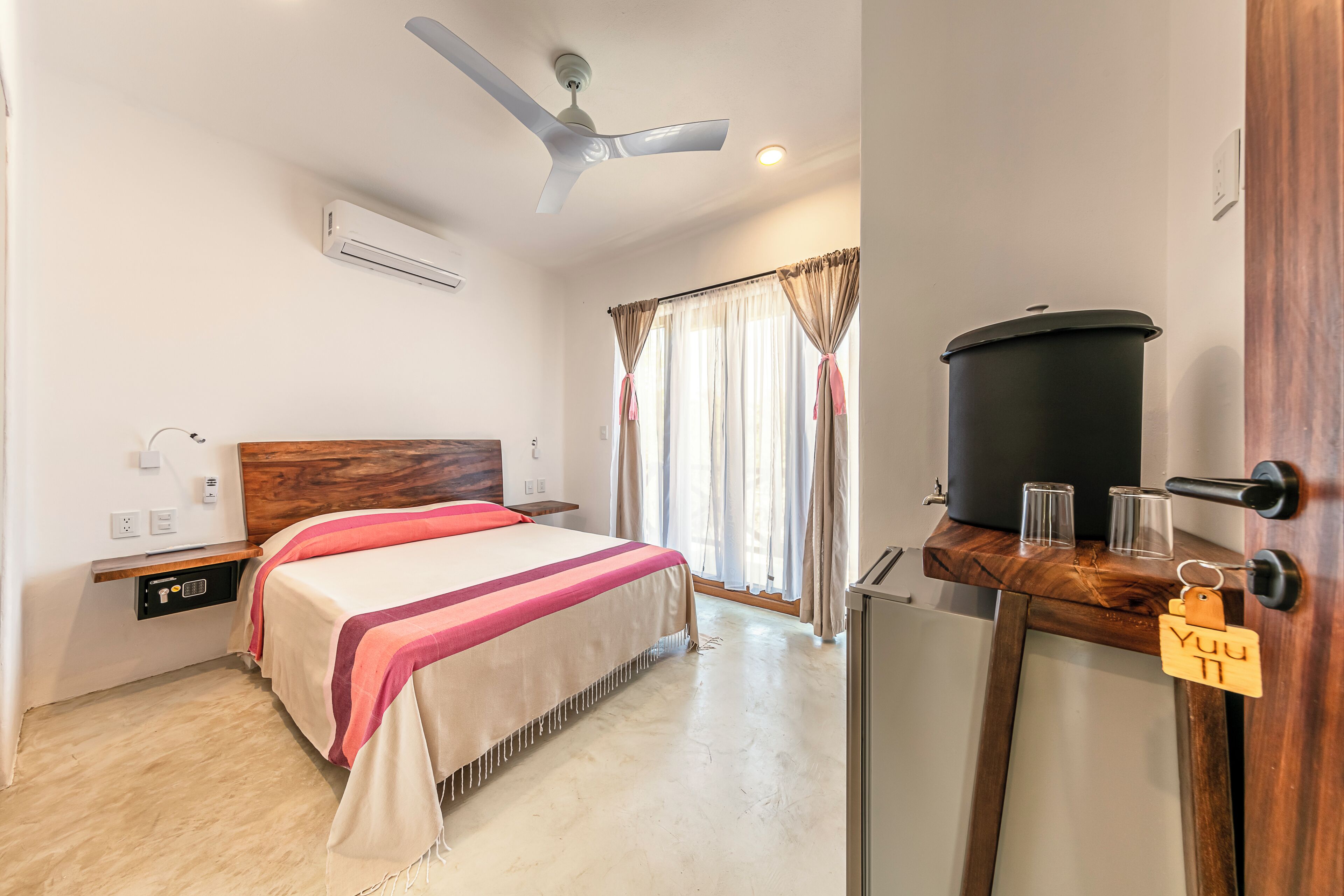Camera d'albergo con un letto, ventilatore a soffitto, condizionatore d'aria, testiera in legno, tende, una finestra, un tavolino con un cestino nero e bicchieri, e una porta con maniglia in legno.