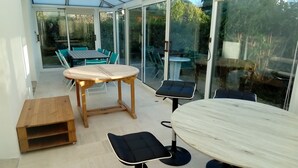 Un patio soleggiato con un tavolo rotondo e sedie, una panca di legno e una porta vetrata che dà su un giardino.