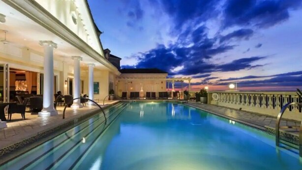 A pool area with a view of the sunset, a covered seating area, and a building with columns.