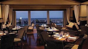 A restaurant with tables and chairs arranged for dining, large windows offering a view of the cityscape, and a warm interior ambiance.