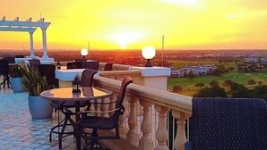 A terrace with a round table and chairs, a pergola, and a view of the sunset over a cityscape.