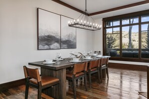 Un coin repas avec une table en bois dressée pour six personnes, un lustre et une grande fenêtre donnant sur les montagnes.