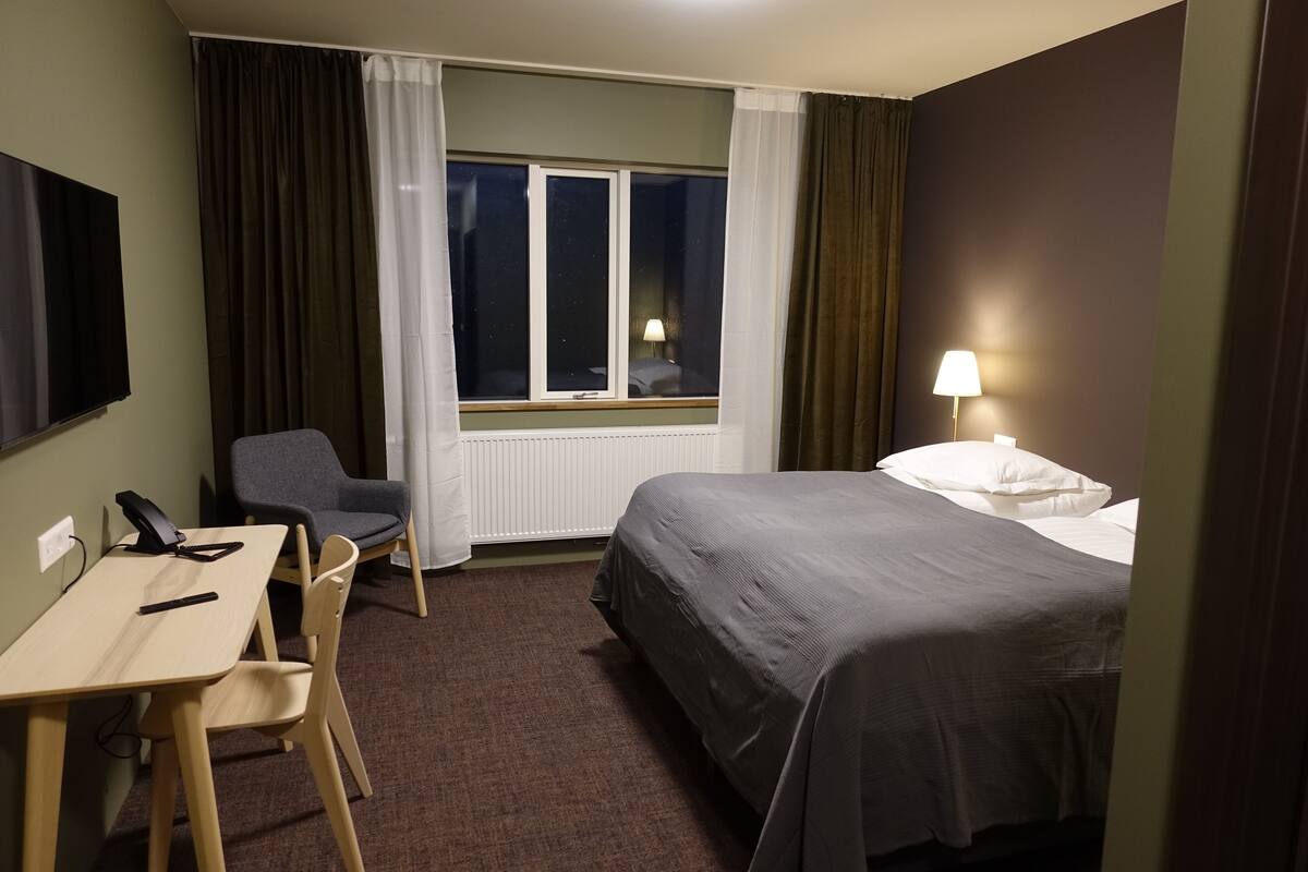 A hotel room with a bed, a desk, a chair, a window with curtains, and a lamp.