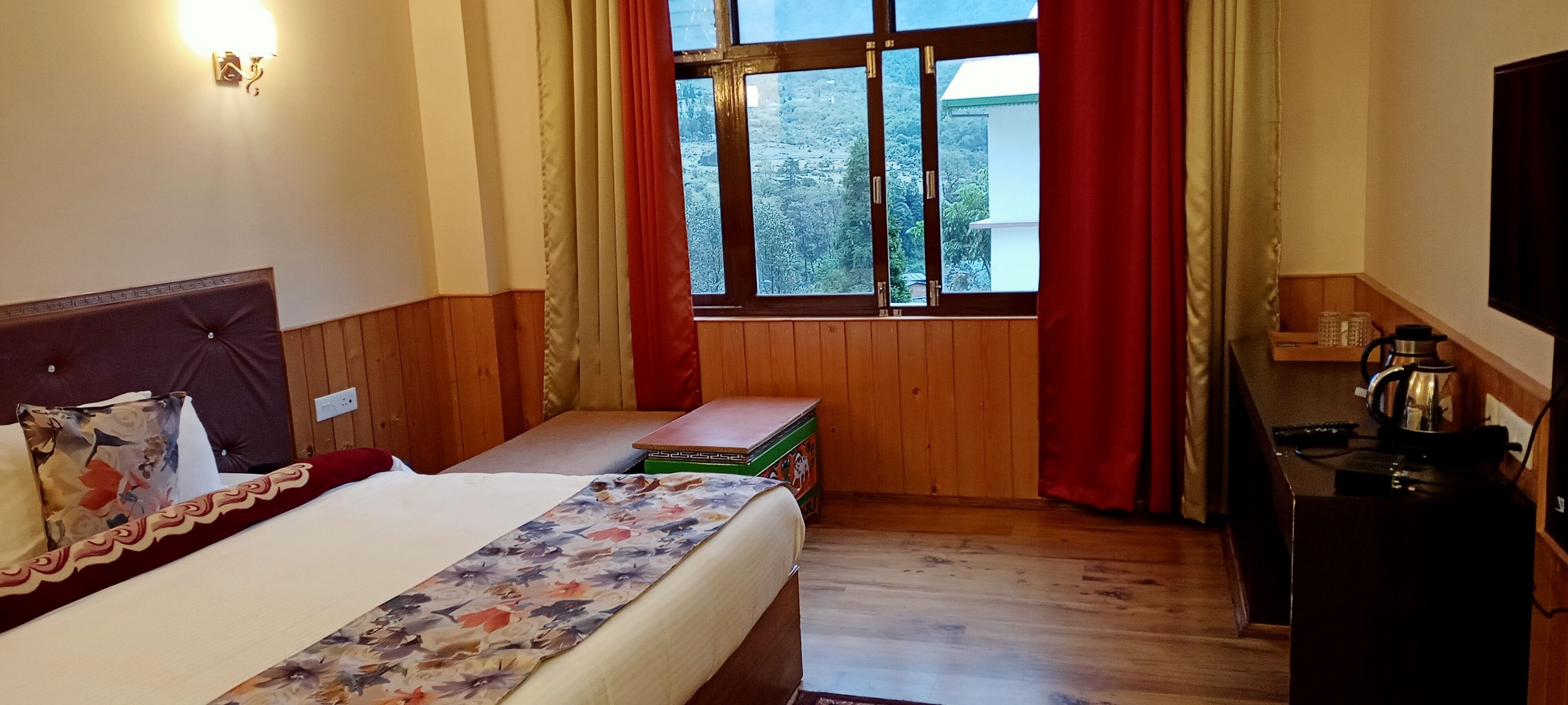 A hotel room with a bed, wooden panel walls, a window with curtains, a small table with a kettle, and a TV on a wooden stand.
