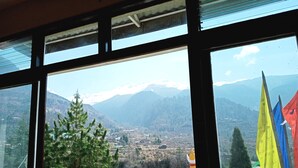 Vista desde una ventana a un paisaje montañoso con picos nevados, un pueblo en la falda de la montaña y coloridas banderas de oración al frente.