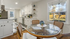 Una cocina moderna con armarios blancos, piso a cuadros blanco y negro, y un comedor con mesa de vidrio y sillas de mimbre.