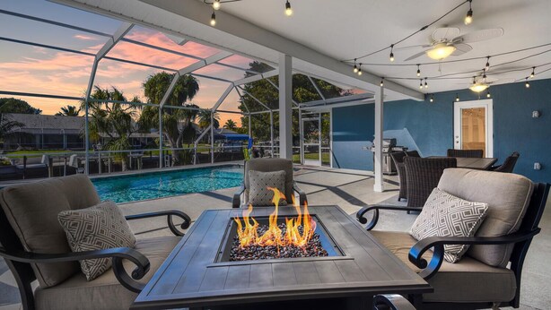 Un patio al aire libre con una chimenea, asientos y vistas a una piscina y edificios al fondo.