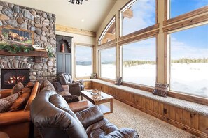 A cozy living room with a stone fireplace, leather sofa, and large windows overlooking a snowy landscape.