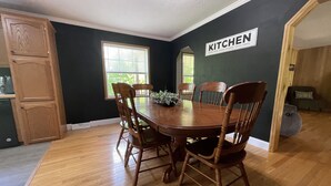 A dining room with a wooden table and chairs, a kitchen sign on the wall, and a window with blinds.