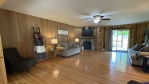 A spacious living room with wooden floors, a fireplace, a ceiling fan, and a television.