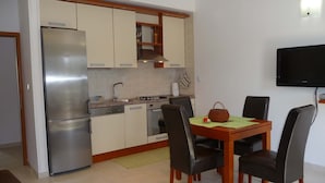 Een kleine keuken met een koelkast, fornuis en wastafel, een eettafel met stoelen, en een flatscreen televisie aan de muur bevestigd.