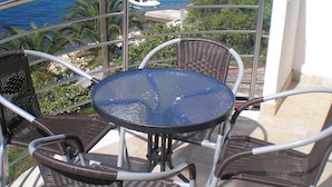 Een balkon met rieten stoelen en een glazen tafel, met uitzicht op een kustgebied met boten en huizen.