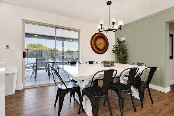 A modern dining area with a large table, chairs, a chandelier, and a clock on the wall.