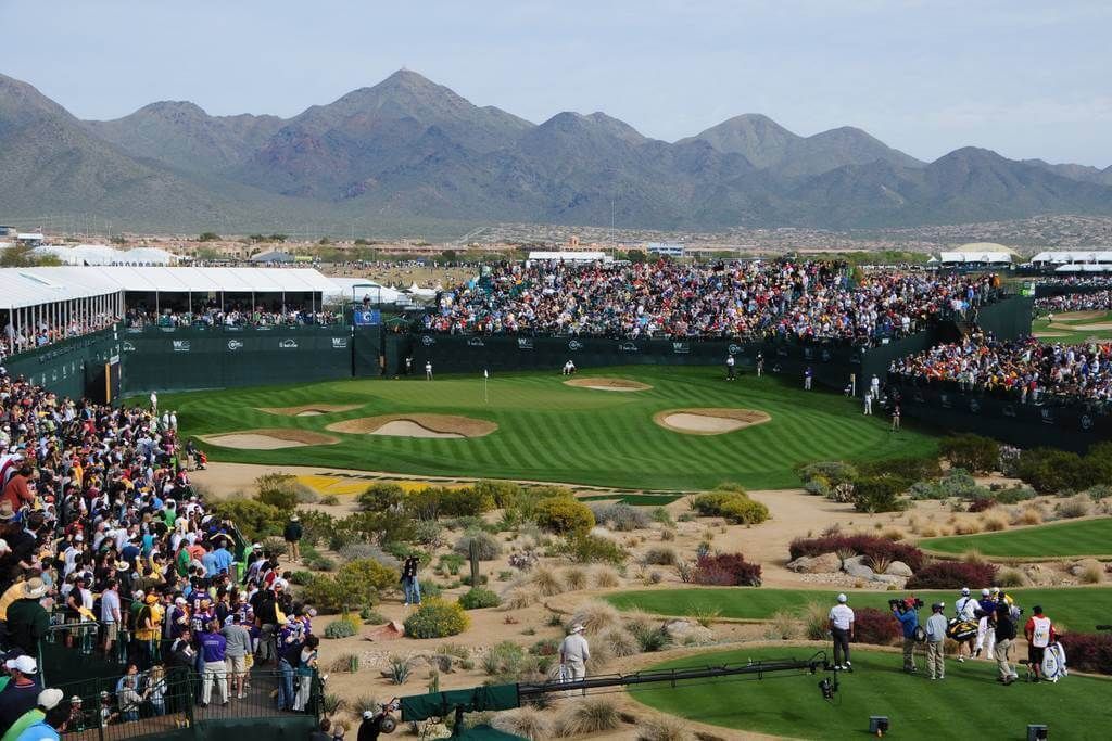 Un campo de golf con espectadores, carpas y montañas al fondo.