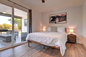 A bedroom with a bed, a nightstand, a lamp, a painting, and a sliding glass door leading to an outdoor area.