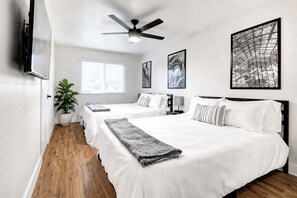 A hotel room with two beds, a ceiling fan, a large window, and artwork on the walls.