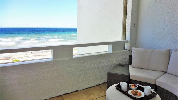 A balcony with a view of the ocean, a white cushioned seating area, and a tray with breakfast items.