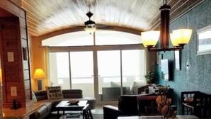 A spacious living area with a high ceiling, a dining table with chairs, a sofa, and a large window providing natural light.