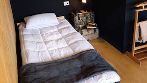 En enkeltseng med et hvidt og gråt dynebetræk, et sengebord med en lampe, og et lille trænatbord med bøger og et glas.