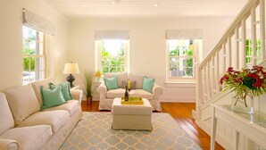 Ein geräumiges Wohnzimmer mit hoher Decke, weißen Wänden und einer Holztreppe. Zwei Sofas mit grünen Kissen, ein Couchtisch mit Flasche und Gläsern sowie ein Blumentopf sind zu sehen.