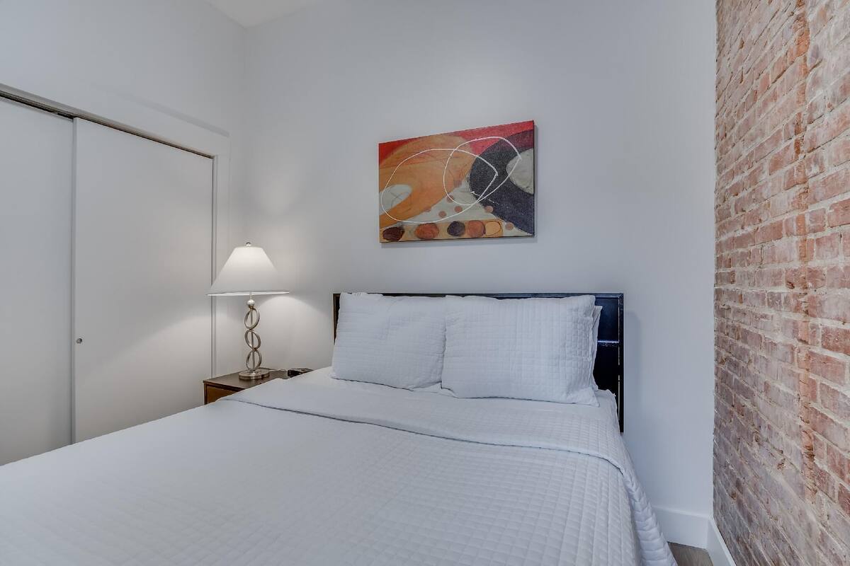 A bedroom with a bed, a nightstand, a lamp, a brick wall, and a painting on the wall.