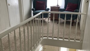 A staircase with white balusters leading to a second-floor living area with two blue armchairs and a red pillow, a wooden side table, and a lamp.