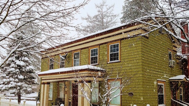A two-story house with a porch, surrounded by a white picket fence, in a snowy setting.