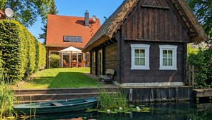 A wooden house with a thatched roof, a pond with lily pads, and a small boat.