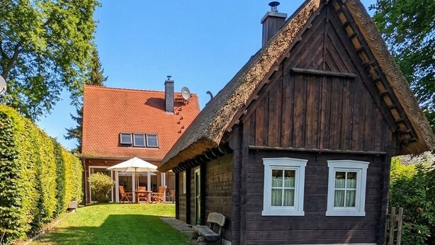 A traditional wooden house with a thatched roof, a pond with ducks, and a clear blue sky.