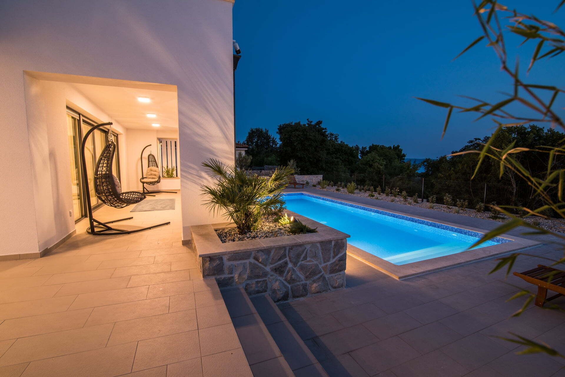A modern outdoor pool area with a stone planter, two black hanging chairs, and a wooden bench.
