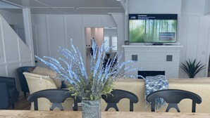 A living room with a wooden dining table, a tall vase with blue flowers, a television mounted on the wall, and a staircase in the background. - BEAUTIFUL LAKE MICHIGAN BEACH HOUSE just a 5-minute walk to the water’s edge! (Muskegon)