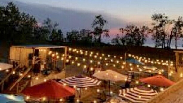 An outdoor seating area with string lights, umbrellas, and a view of trees and a building in the background. - BEAUTIFUL LAKE MICHIGAN BEACH HOUSE just a 5-minute walk to the water’s edge! (Muskegon)