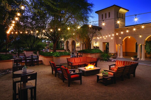 Relax in style with a sofa, chairs, and table surrounding a central fire feature, amidst lush greenery and string lights.