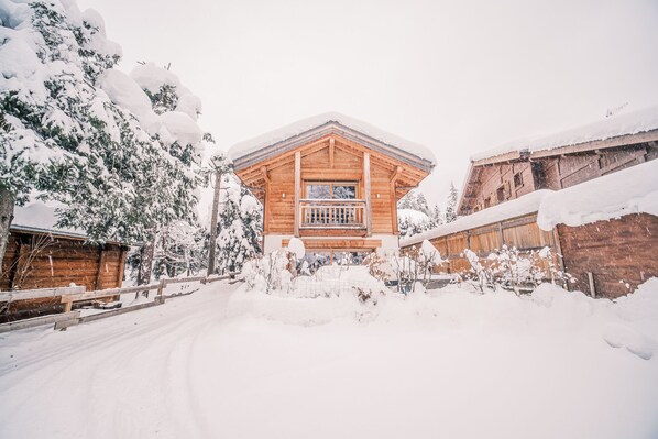 A wooden cabin with a balcony, surrounded by snow-covered trees and a wooden fence.