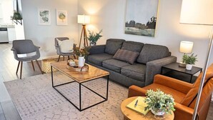 Uma sala de estar moderna com um sofá cinzento, uma mesa de centro de madeira, uma mesa redonda com uma cadeira e uma mesinha lateral com uma luminária.