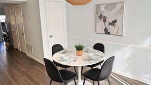 Uma área de refeições com uma mesa redonda posta para quatro pessoas, cadeiras pretas, uma luminária pendente trançada e uma pintura abstrata moderna na parede.