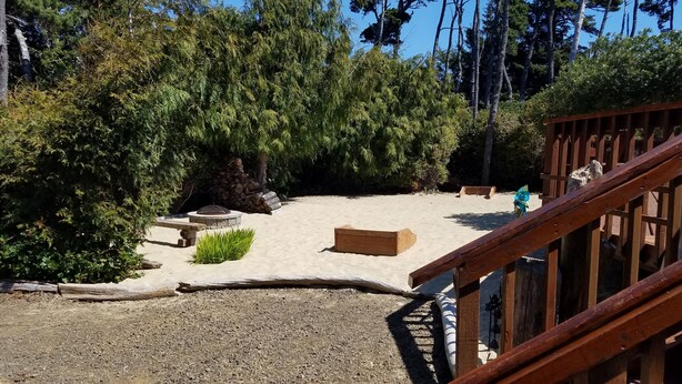 Un espacio al aire libre con arena, un hogar de piedra, bancos de madera y una terraza de madera.