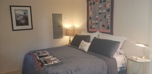 A neatly made bed with a grey blanket and pillows, a bedside lamp, a framed picture on the wall, and a quilt with a nautical theme.