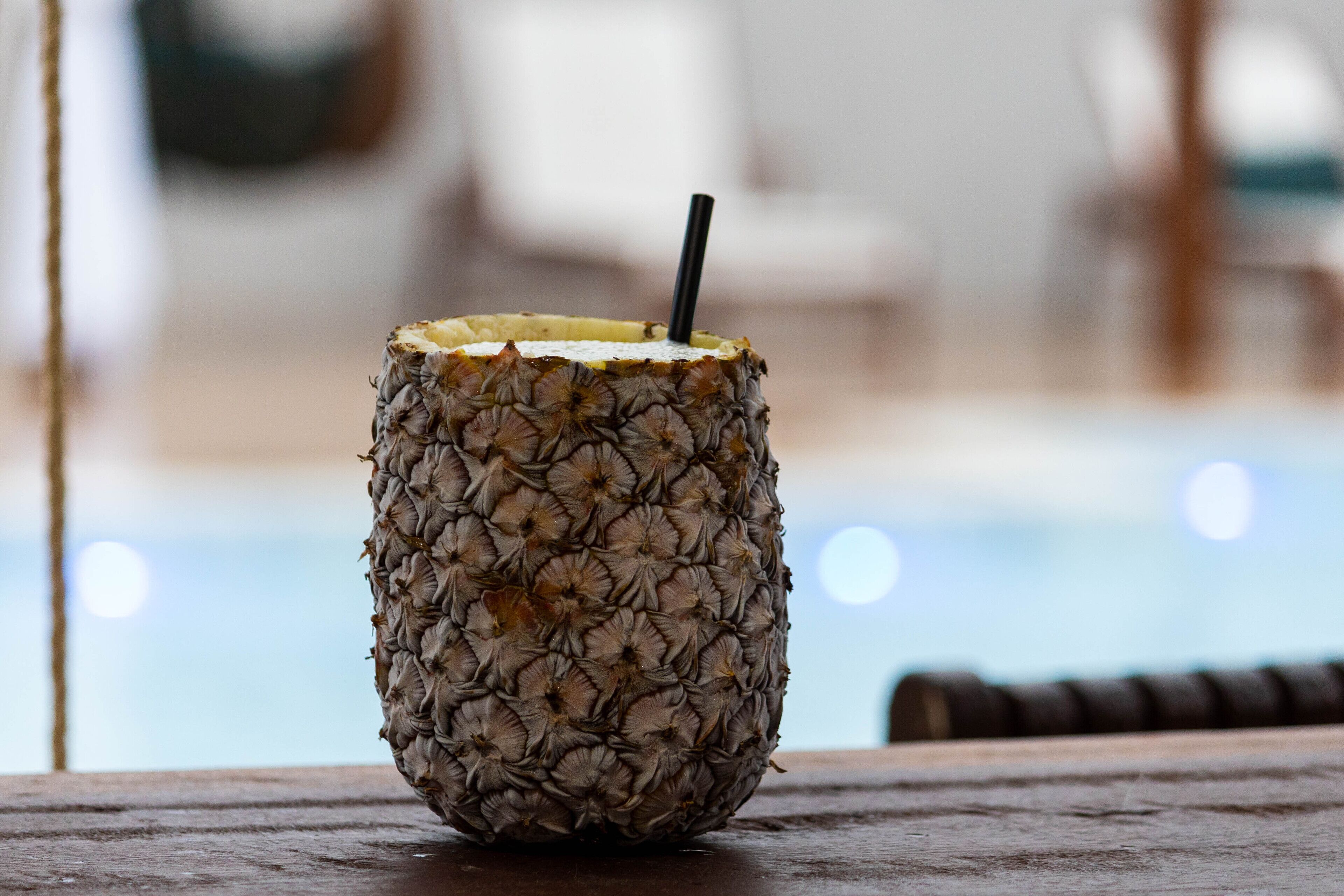 Un cocktail all'ananas con una cannuccia in un bicchiere, appoggiato su un piano di legno vicino a una piscina.