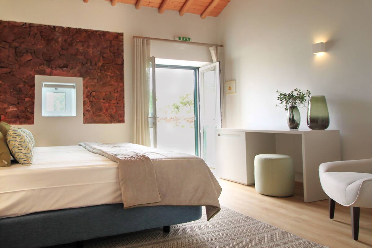 Un dormitorio con cama, escritorio, silla, taburete pequeño e iluminación montada en la pared.