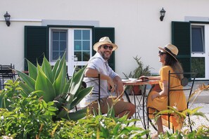Dos personas sentadas en una mesa al aire libre, con vista a un edificio de fachada blanca y contraventanas verdes.