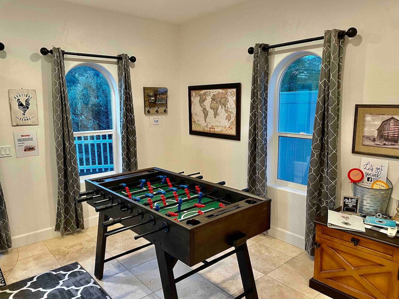 A room with a foosball table, two windows with curtains, a map, and a small cabinet with a sign.