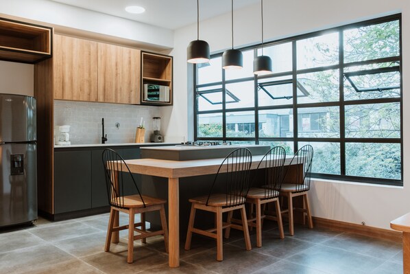 Una cocina moderna con armarios de madera, un refrigerador de acero inoxidable, una isla grande con sillas y un ventanal con vista a árboles.