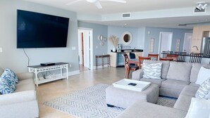 A modern living room with a flat-screen TV, grey sofas, a coffee table, and a dining area with chairs and a small kitchen. - DOWN BY THE BAY! Gulf Facing unit! ASK ABOUT DISCOUNTS!! (Orange Beach)