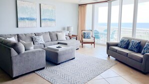 A spacious living room with a gray sofa, a coffee table, and a view of the city through large windows. - DOWN BY THE BAY! Gulf Facing unit! ASK ABOUT DISCOUNTS!! (Orange Beach)
