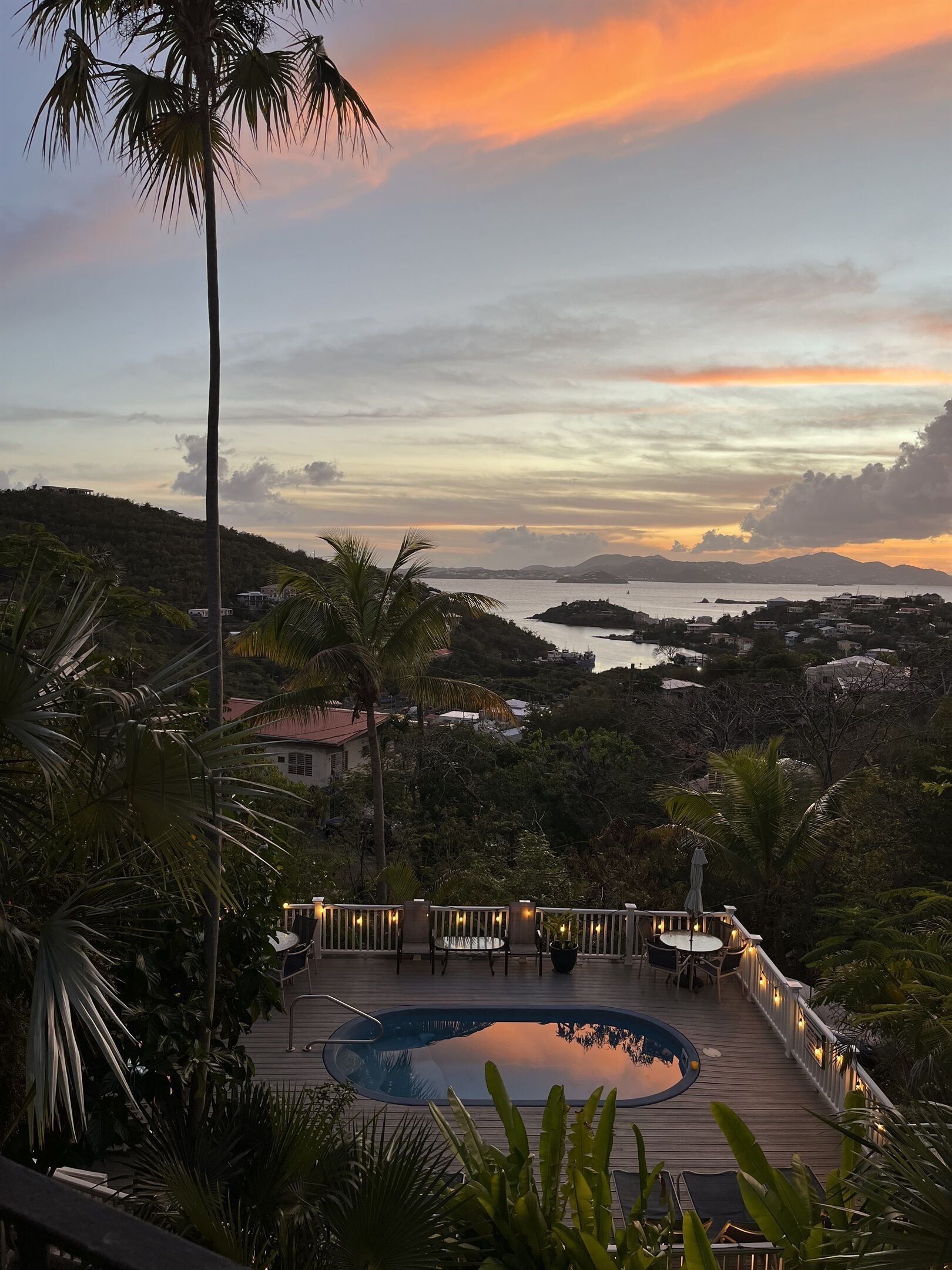 A pool area with a wooden deck, surrounded by palm trees and a view of a coastal landscape during sunset.