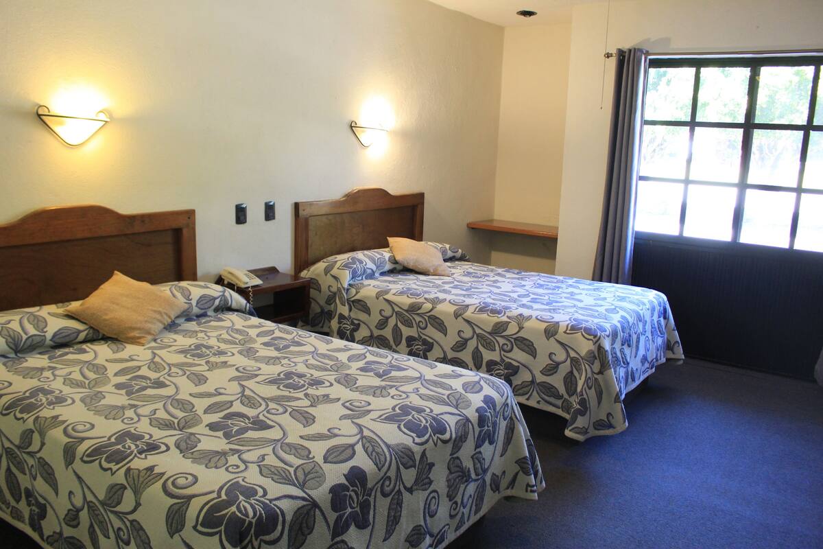 A hotel room with two beds, each with a floral-patterned bedspread, bedside lamps, and a window with curtains.