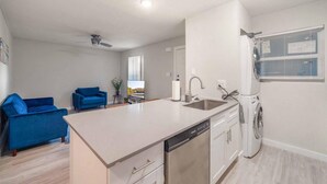 A modern kitchen with a dishwasher, sink, and a window with blinds. There is a blue sofa in the living area. - Stylish 2BR Near UT Hyde Park Evonify (Austin)